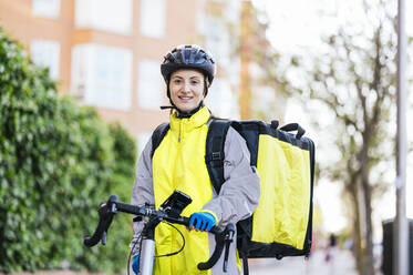 Junge Frau mit Thermotasche steht mit dem Fahrrad beim Ausliefern von Lebensmitteln auf einer Straße in der Stadt und schaut in die Kamera - ADSF28420