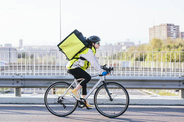 Seitenansicht eines weiblichen Kuriers mit Thermotasche, der mit dem Fahrrad auf einer Brücke fährt, während er Lebensmittel in der Stadt ausliefert - ADSF28423