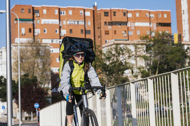 Fröhlicher weiblicher Kurier mit Thermotasche fährt mit dem Fahrrad auf einer Brücke, während er Lebensmittel in der Stadt ausliefert - ADSF28424