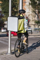 Rückenansicht einer Lieferfrau mit leerer Thermotasche, die wegschaut und auf einem Fahrrad auf dem Bürgersteig in der Nähe einer Straße in der Stadt sitzt - ADSF28426