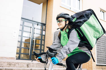 Niedriger Winkel der weiblichen Kurier mit thermischen Tasche lächelnd und Radfahren auf der Straße während der Auslieferung auf sonnigen Tag in der Stadt - ADSF28440