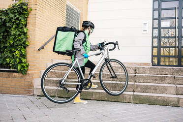 Weiblicher Ganzkörperkurier mit Thermotasche, der sein Fahrrad auf der Treppe zum Gebäudeeingang trägt, während er eine Lieferung macht - ADSF28441