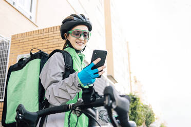 Junge, fröhliche Frau mit Thermotasche, die auf dem Fahrrad sitzend ihr Smartphone mit einer GPS-Karte durchsucht, um Lebensmittel in der Stadt auszuliefern - ADSF28445