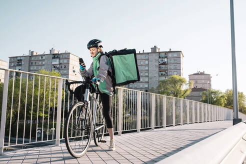 Ganzkörper junger weiblicher Kurier in Uniform und Helm auf dem Fahrrad sitzend und auf einer Brücke in der Stadt eine GPS-Karte auf dem Handy durchsuchend - ADSF28453