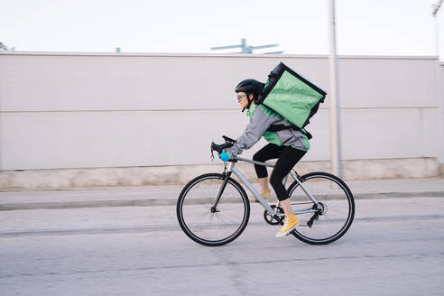 Seitenansicht einer weiblichen Kurierin mit Thermotasche, die mit dem Fahrrad auf der Straße unterwegs ist und Lebensmittel in der Stadt ausliefert, Bewegungsunschärfe - ADSF28454