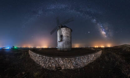 Picturesque scenery of Milky Way in dark night sky above aged stone windmill tower with glowing lights in distance - ADSF28472