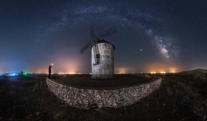 Picturesque scenery of Milky Way in dark night sky above aged stone windmill tower with glowing lights in distance - ADSF28473