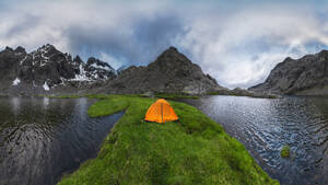 Orangefarbenes Zelt am grasbewachsenen Ufer des Sees Laguna Grande vor der Bergkette Sierra de Gredos und dem bewölkten Himmel in Avila, Spanien - ADSF28480