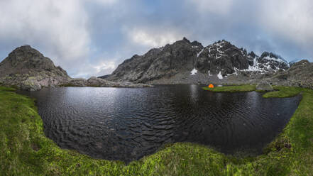 Orangefarbenes Zelt am grasbewachsenen Ufer des Sees Laguna Grande vor der Bergkette Sierra de Gredos und dem bewölkten Himmel in Avila, Spanien - ADSF28481