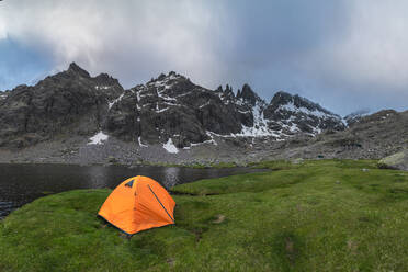 Orangefarbenes Zelt am grasbewachsenen Ufer des Sees Laguna Grande vor der Bergkette Sierra de Gredos und dem bewölkten Himmel in Avila, Spanien - ADSF28483