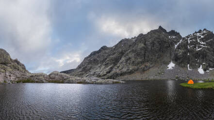 Orangefarbenes Zelt am grasbewachsenen Ufer des Sees Laguna Grande vor der Bergkette Sierra de Gredos und dem bewölkten Himmel in Avila, Spanien - ADSF28485