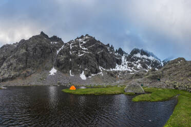 Orangefarbenes Zelt am grasbewachsenen Ufer des Sees Laguna Grande vor der Bergkette Sierra de Gredos und dem bewölkten Himmel in Avila, Spanien - ADSF28486