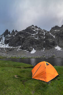 Orangefarbenes Zelt am grasbewachsenen Ufer des Sees Laguna Grande vor der Bergkette Sierra de Gredos und dem bewölkten Himmel in Avila, Spanien - ADSF28487