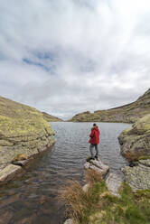 Side view of anonymous man in outerwear standing on stones near Laguna Grande lake amidst mountains of Sierra de Gredos range in Avila, Spain - ADSF28491