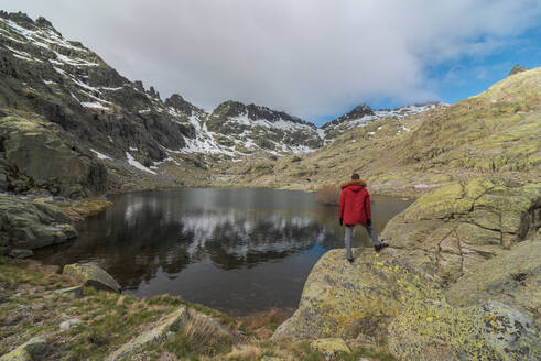 Rückenansicht eines anonymen Mannes in Oberbekleidung, der auf Steinen in der Nähe des Sees Laguna Grande inmitten der Berge der Sierra de Gredos in Avila, Spanien, steht - ADSF28492