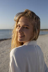 Blonde Frau mit langen Haaren steht am Strand und schaut in die Kamera - ADSF28508