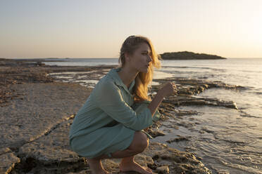 Long-haired blonde woman crouched down on the beach looking into the distance - ADSF28511
