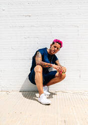Selbstbewusster queerer Mann mit bunten Nägeln und rosa Haaren, der im Sommer auf der Straße sitzt und in die Kamera schaut - ADSF28535