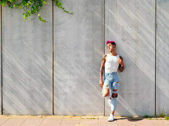 Trendy gay male with pink hair and ripped jeans leaning on wall in city and looking away - ADSF28536