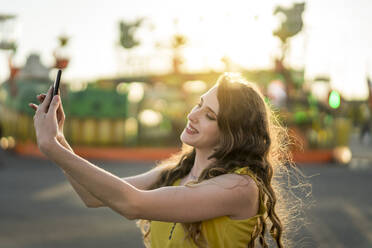 Content female taking self portrait on mobile phone while having fun in amusement park in evening in summer - ADSF28561