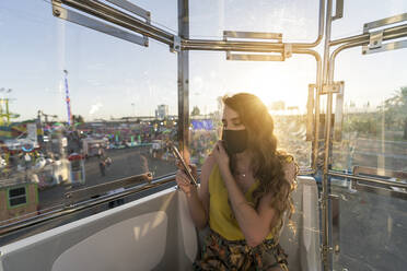 Female in protective mask sitting in cabin of Ferris wheel and browsing mobile phone during ride while having fun at fairground during coronavirus - ADSF28562