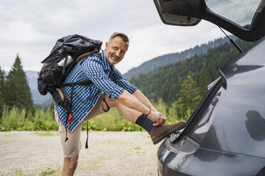 Männlicher Rucksacktourist bindet Schnürsenkel, während er sich an den Kofferraum eines Autos lehnt - DIGF16306