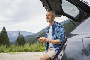 Nachdenklicher Mann hält sein Smartphone im Kofferraum eines Autos - DIGF16309