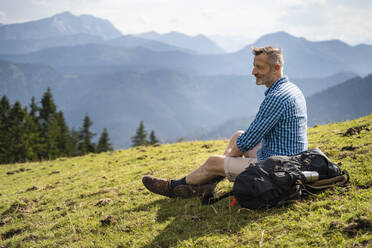 Mature man looking at mountain while sitting by backpack - DIGF16343