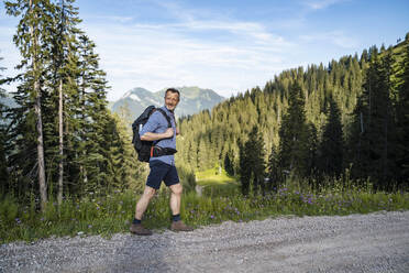 Male hiker with backpack looking over shoulder while walking on dirt road - DIGF16380