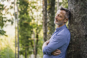 Älterer Mann mit geschlossenen Augen lehnt an einem Baum im Wald - DIGF16390
