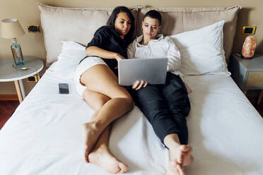 Junge queere Freundinnen benutzen einen Laptop, während sie zu Hause auf dem Bett liegen - MEUF04076