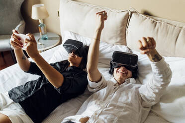 Young gay women enjoying virtual reality while lying on bed at home - MEUF04085