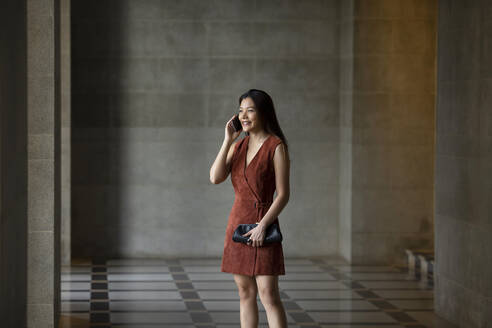 Smiling woman talking on phone while standing at corridor - EAF00047