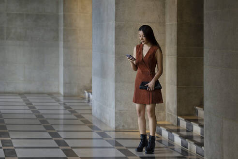 Woman with clutch bag using phone while standing inside passage - EAF00050