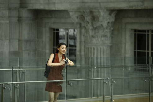 Woman holding camera while leaning on glass railing - EAF00051