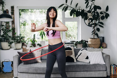 Smiling woman exercising with plastic hoop in living room - ASGF01103