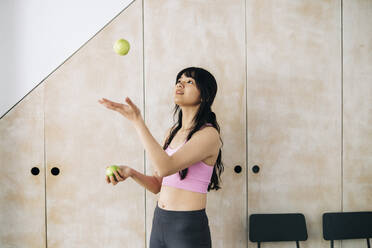 Woman juggling apples while standing in front of cabinet at home - ASGF01132