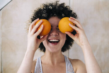 Happy woman covering eyes with oranges at home - ASGF01136