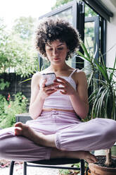 Young woman using mobile phone while sitting cross-legged on chair - ASGF01137