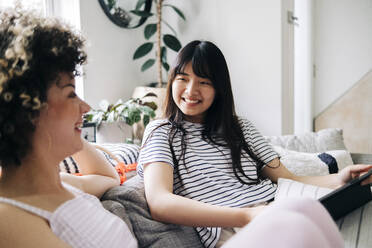 Happy woman with digital tablet looking at friend while sitting on sofa - ASGF01142