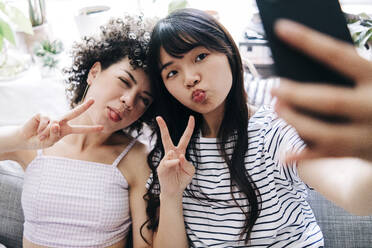 Female friends showing peace sign while taking selfie through smart phone at home - ASGF01148