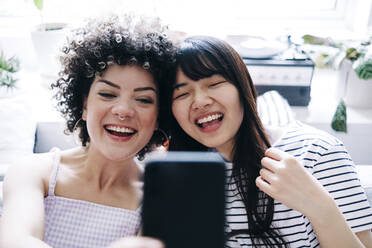Cheerful female friends taking selfie through smart phone at home - ASGF01149
