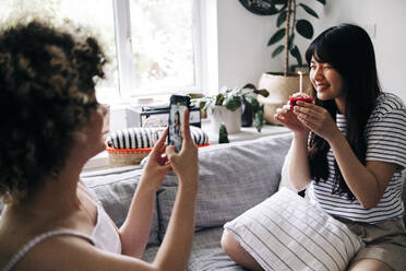 Woman taking photo of friend holding cupcake through mobile phone at home - ASGF01150