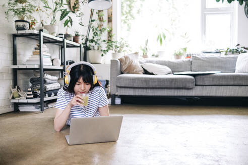 Junge Frau mit Kopfhörern, die einen Smoothie trinkt, während sie zu Hause einen Laptop benutzt - ASGF01167