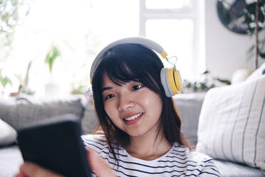 Lächelnde Frau mit drahtlosen Kopfhörern, die zu Hause ein Smartphone benutzt - ASGF01170