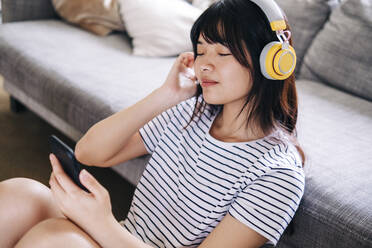 Woman with mobile phone listening music through headphones in living room - ASGF01181