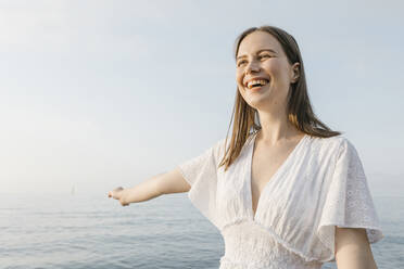 Beautiful young woman laughing while standing with arm outstretched at beach - XLGF02207