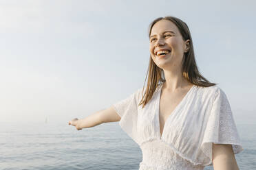 Schöne junge Frau, die lachend mit ausgestrecktem Arm am Strand steht - XLGF02207