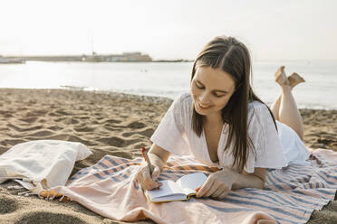 Smiling woman writing in book while lying on towel at beach - XLGF02217