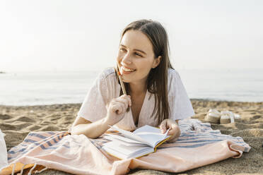 Nachdenkliche glückliche Frau, die einen Stift hält, während sie sich am Strand ausruht - XLGF02218